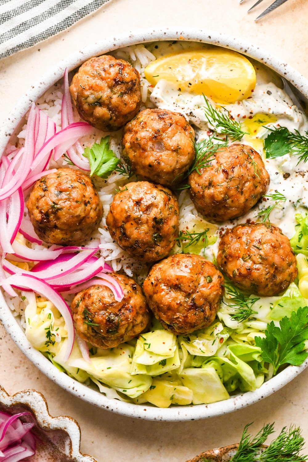 Baked Greek chicken meatballs in a shallow bowl with rice, cabbage salad, thinly sliced pickled red onions, creamy dairy free tzatziki sauce, and a lemon wedge. Bowl is garnished with scattered fresh dill and parsley, black pepper, and a tiny bit of aleppo chili flakes.