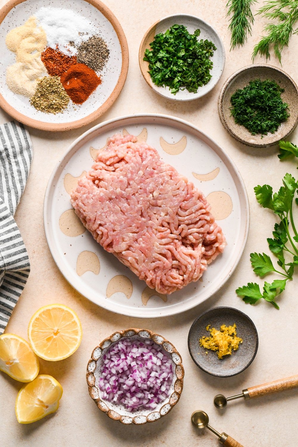 Ingredients for making Greek chicken meatballs in various ceramics, on a cream colored background with a striped linen and sprigs of fresh dill and parsley peeking into the frame. 