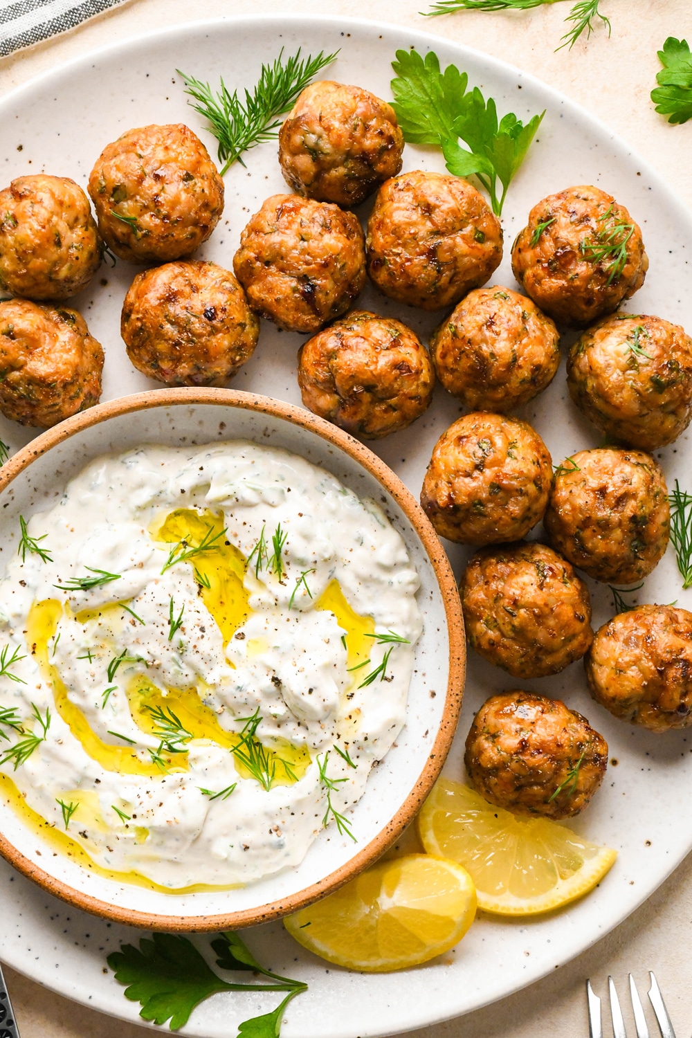 Baked Greek chicken meatballs on a serving platter, next to a small dish of tzatziki, and garnished with a few sprigs of fresh dill and fresh parsley.