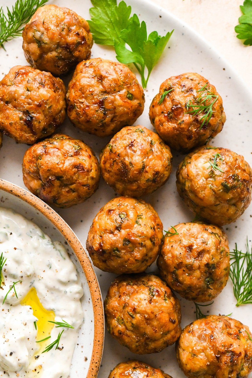 Baked Greek chicken meatballs on a serving platter, next to a small dish of tzatziki, and garnished with a few sprigs of fresh dill and fresh parsley.