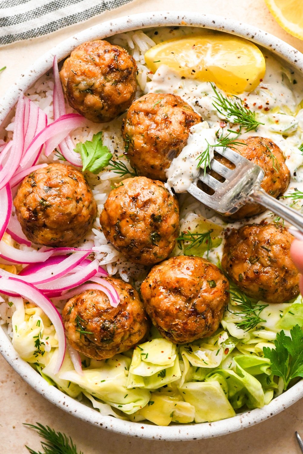 Baked Greek chicken meatballs in a shallow bowl with rice, cabbage salad, thinly sliced pickled red onions, creamy dairy free tzatziki sauce, and a lemon wedge. Bowl is garnished with scattered fresh dill and parsley, black pepper, and a tiny bit of aleppo chili flakes. A fork that's been dipped in the tzatziki is spearing one of the meatballs to lift out for a bite.