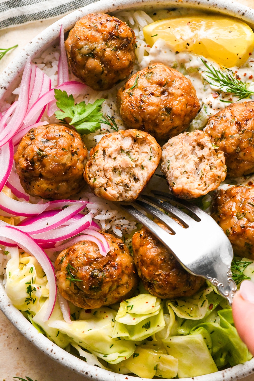 Baked Greek chicken meatballs in a shallow bowl with rice, cabbage salad, thinly sliced pickled red onions, creamy dairy free tzatziki sauce, and a lemon wedge. Bowl is garnished with scattered fresh dill and parsley, black pepper, and a tiny bit of aleppo chili flakes. One of the meatballs is cut in half to show the interior texture, and a fork is angled into the bowl near the cut meatball.