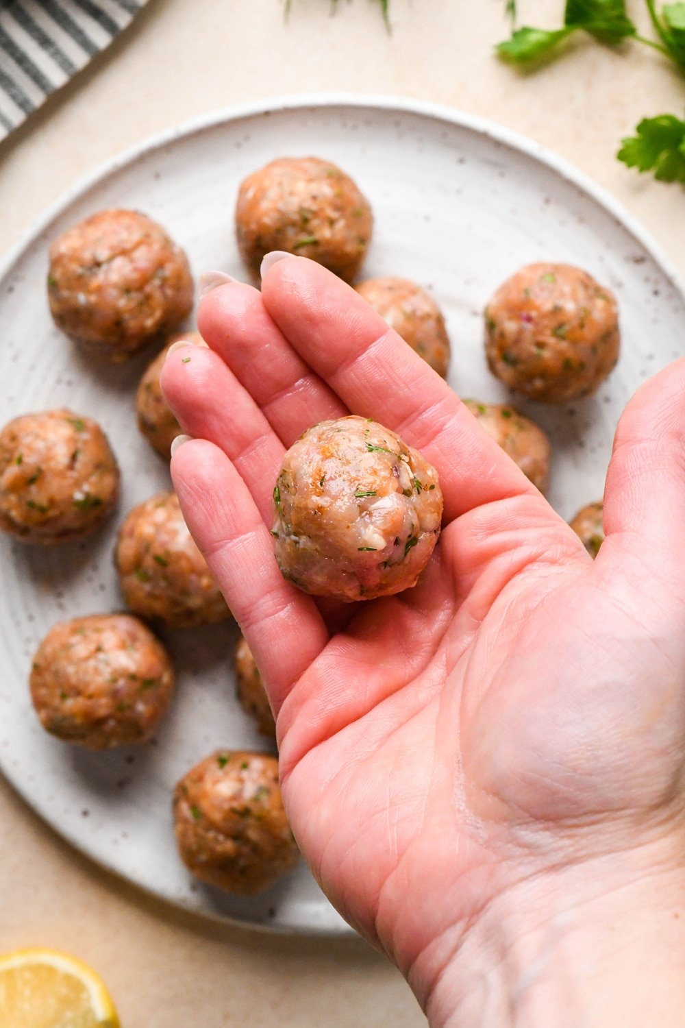 How to make Greek chicken meatballs: A hand that has been wet with water showing a meatball after rolling it to shape over a plate of meatballs that have already been shaped.