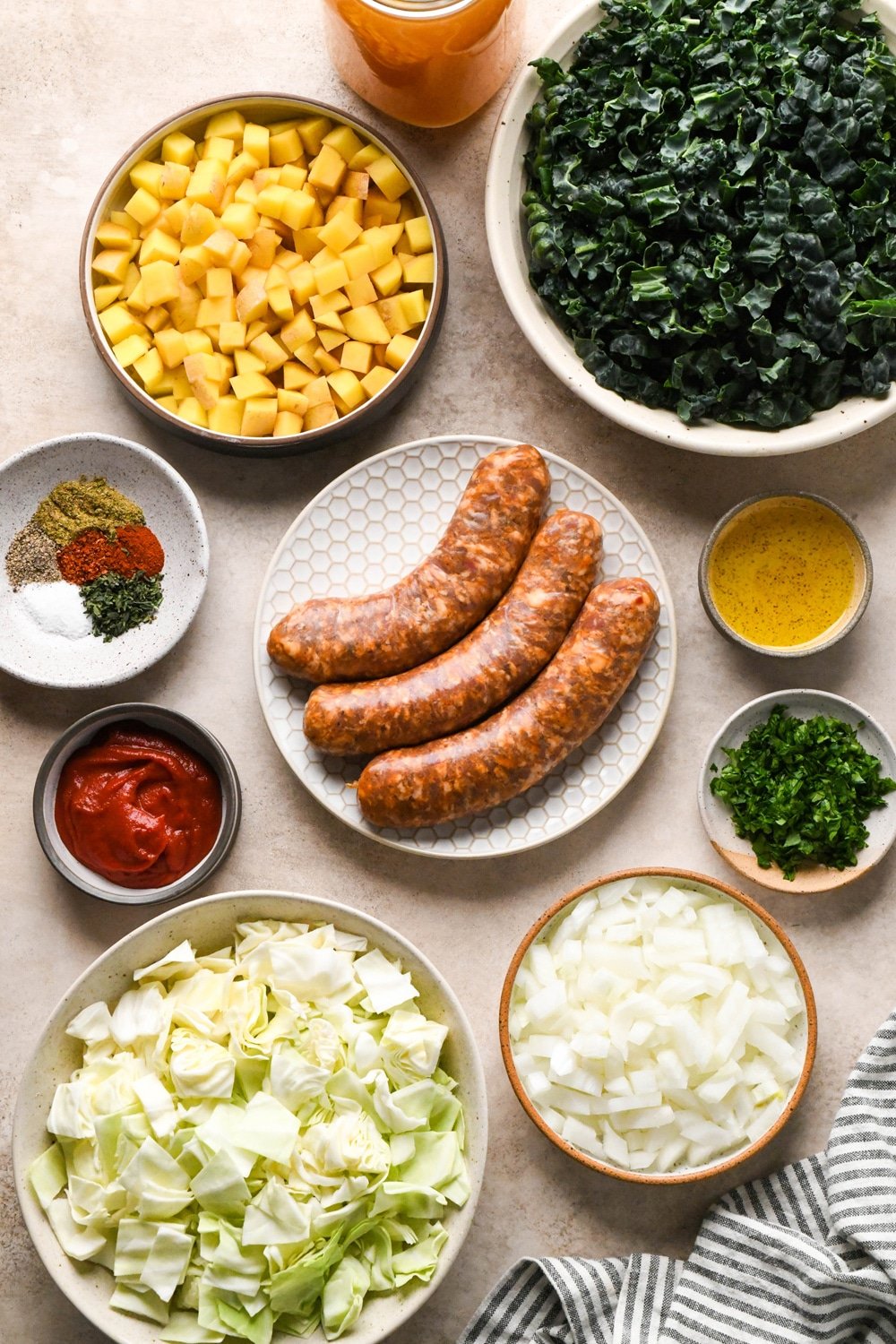 Ingredients for Brothy Sausage and Potato Soup with Kale and Cabbage in various ceramics on a light creamy brown background