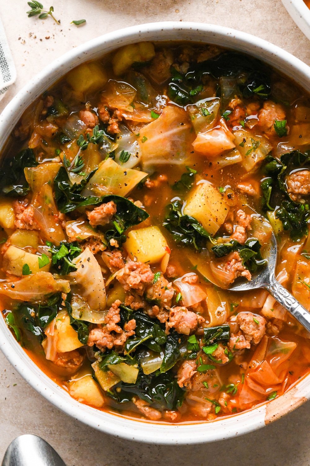 A bowl of brothy sausage and potato soup with a spoon angled into the broth and lifting out a bite.