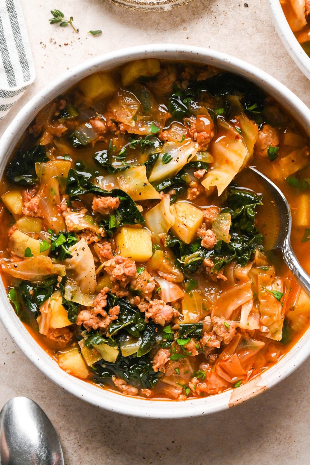 A bowl of brothy sausage and potato soup with a spoon dipping into the right side of the bowl.