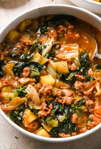 A bowl of brothy sausage and potato soup with a spoon angled into the broth.