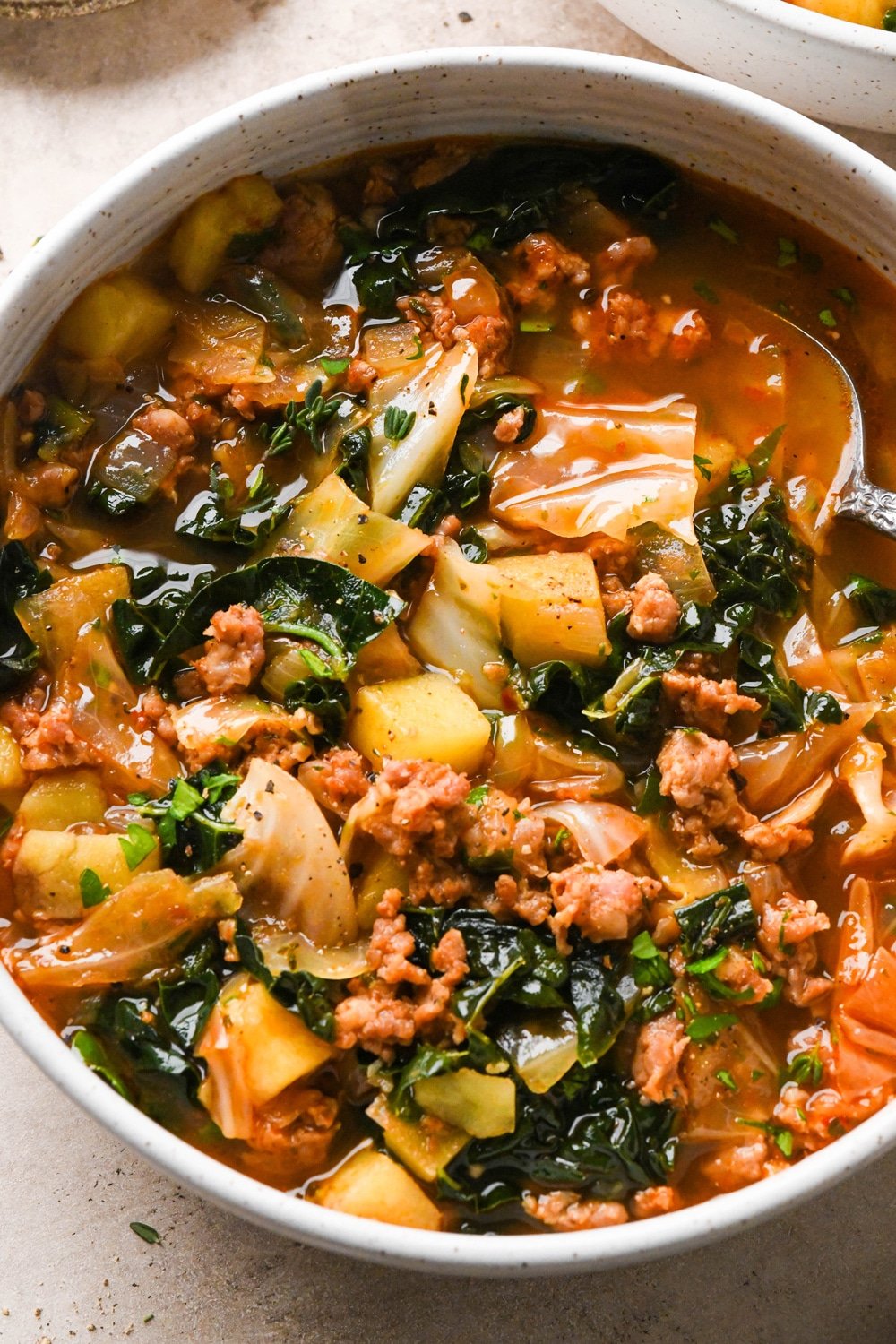 A bowl of brothy sausage and potato soup with a spoon angled into the broth. 