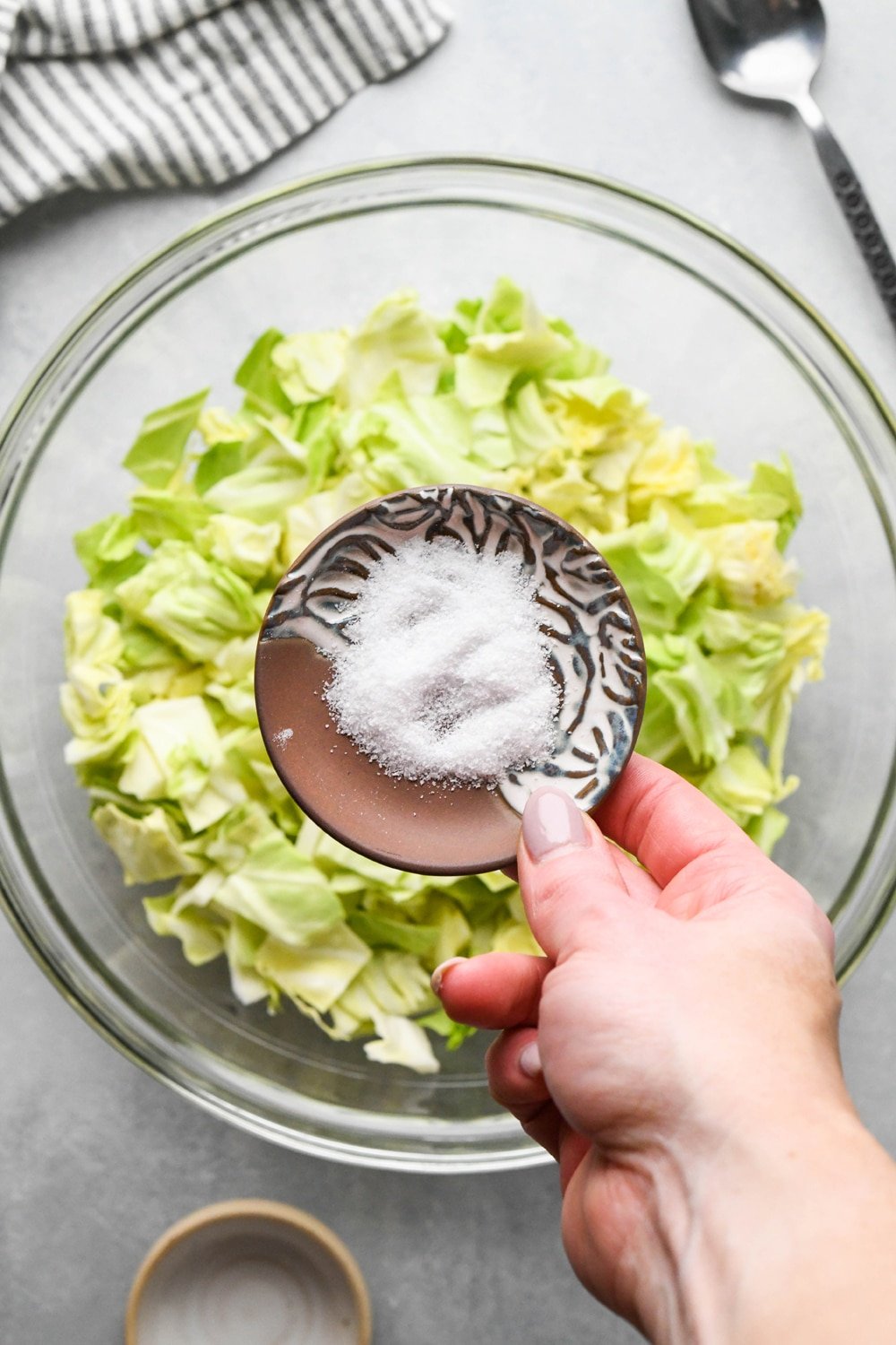 How to make chopped green cabbage salad: A hand adding kosher salt from a small ceramic dish to the bowl of cabbage. 