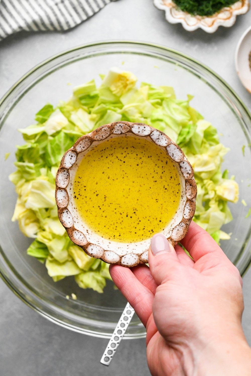 How to make chopped green cabbage salad: A hand adding olive oil from a small ceramic dish to the bowl of cabbage.