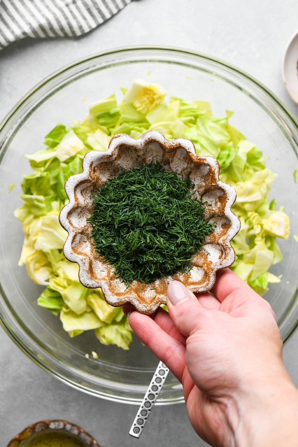 How to make chopped green cabbage salad: A hand adding fresh chopped dill from a small ceramic dish to the bowl of cabbage.