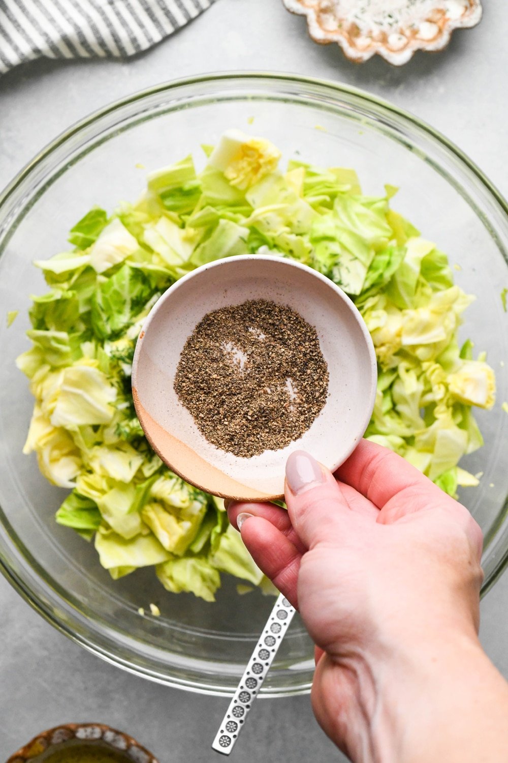 How to make chopped green cabbage salad: A hand adding ground black pepper from a small ceramic dish to the bowl of cabbage.