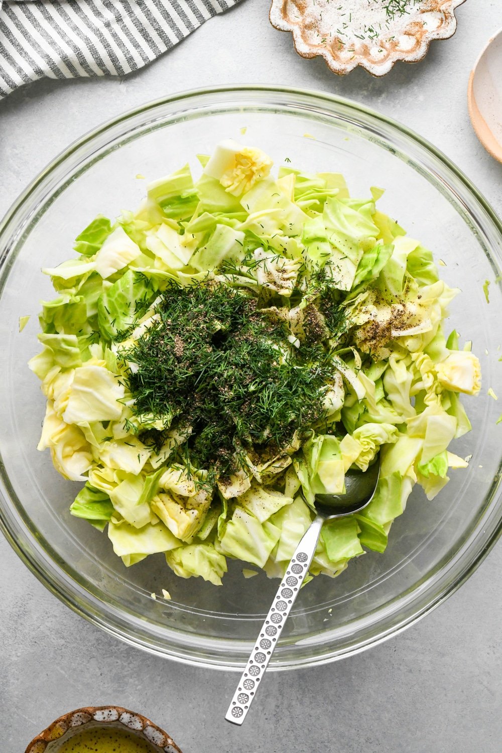 How to make chopped green cabbage salad: Chopped cabbage in glass bowl with olive oil, fresh dill, and black pepper on top before mixing.