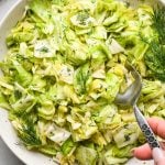 Chopped green cabbage salad with fresh dill in a large shallow serving bowl, garnished with extra sprigs of fresh dill and a spoon is dipping into the salad to lift out a portion for serving.