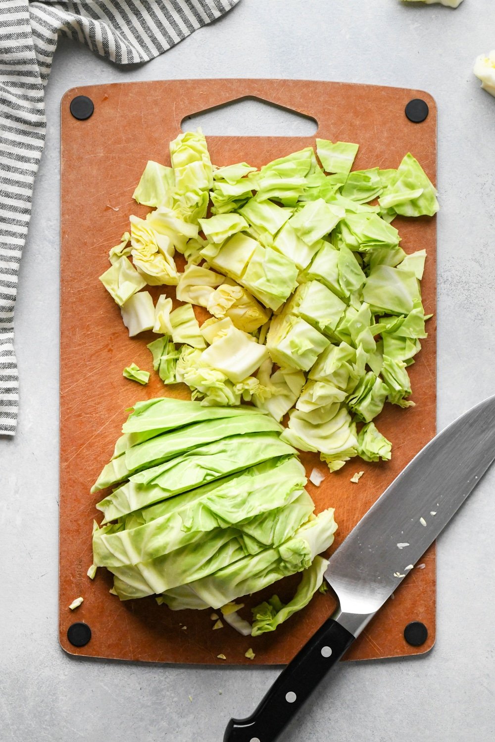 How to make chopped green cabbage salad: Step-by-step showing how to roughly chop cabbage. Small head of green cabbage is cut in half on a cutting board, facing flat side down and cabbage is cut into half inch thick strips. One half of the cabbage has been chopped into bite sized pieces.