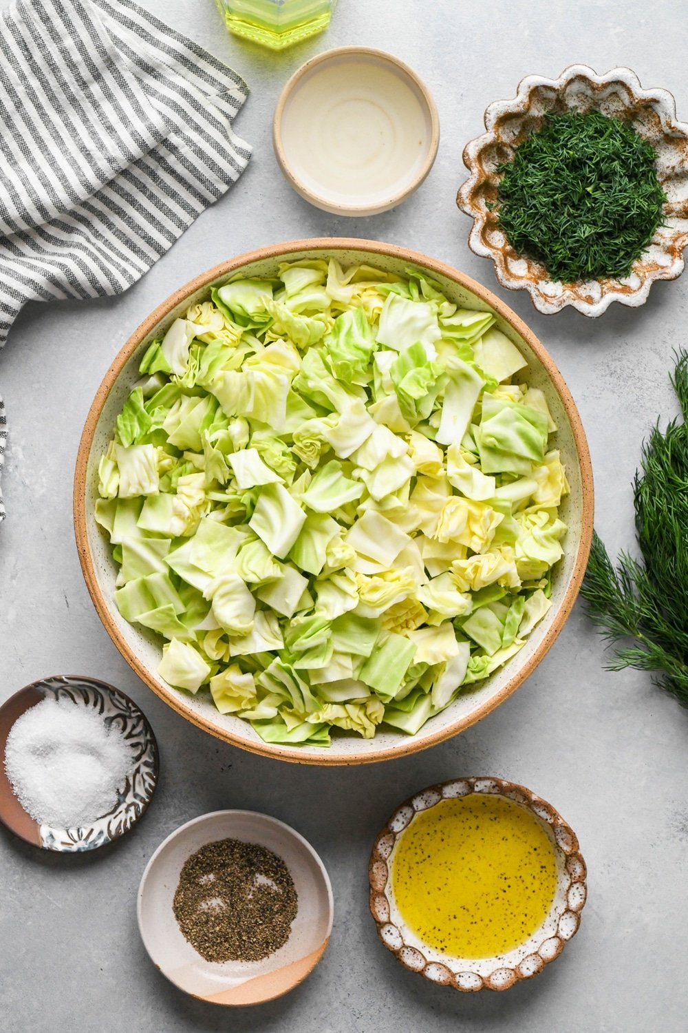 Ingredients for chopped green cabbage salad in various ceramic dishes on a light grey colored background with a striped navy and cream linen peeking into the frame.