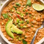 A hearty-looking bowl of creamy dairy free buffalo chicken chili garnished with sliced avocado, thinly sliced green onions, and chopped cilantro, with a spoon and lime wedge angled into the bowl.