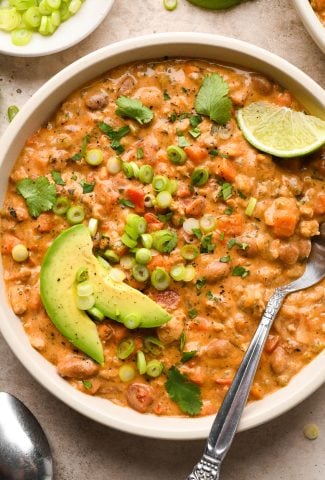 A hearty-looking bowl of creamy dairy free buffalo chicken chili garnished with sliced avocado, thinly sliced green onions, and chopped cilantro, with a spoon and lime wedge angled into the bowl.