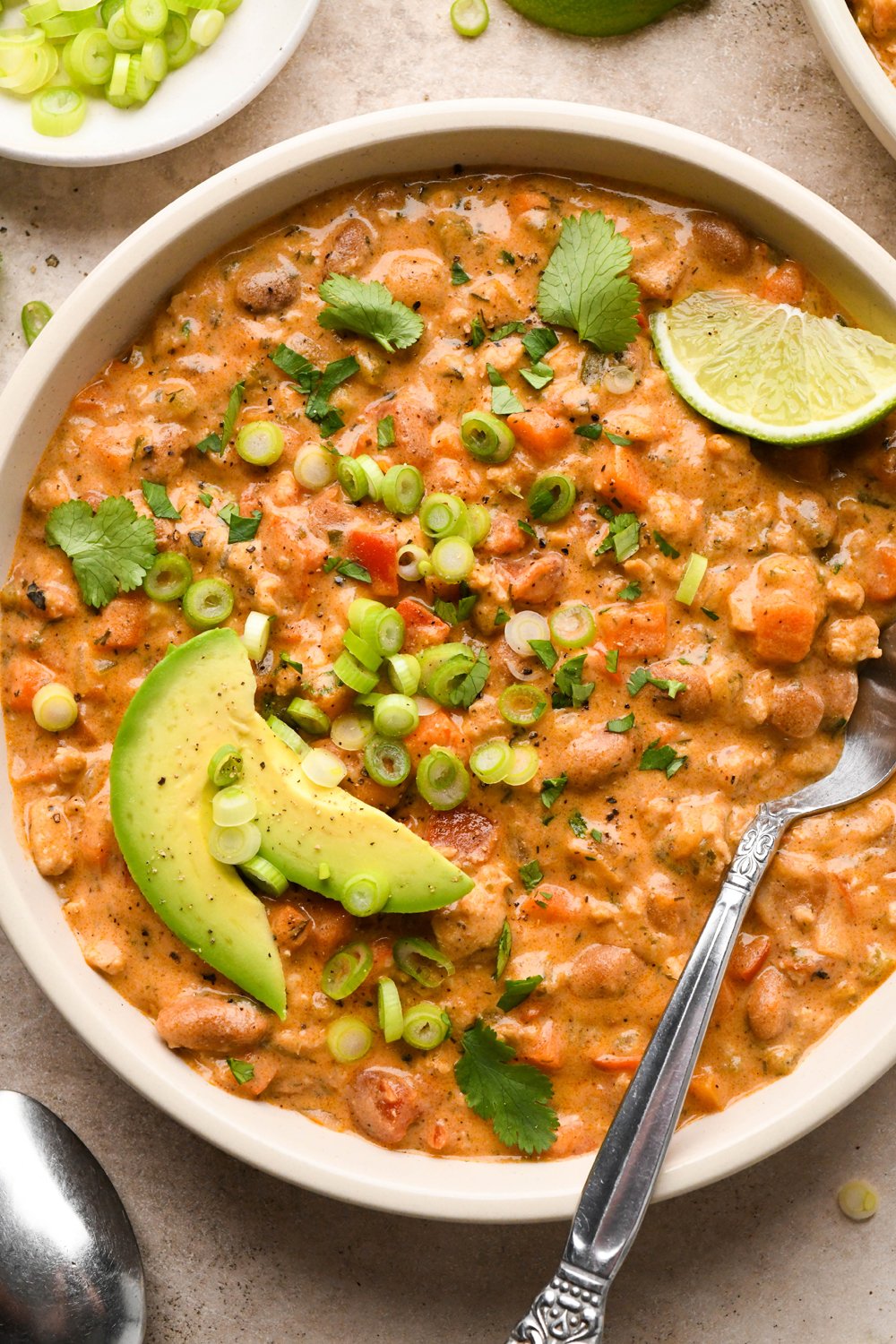 A hearty-looking bowl of creamy dairy free buffalo chicken chili garnished with sliced avocado, thinly sliced green onions, and chopped cilantro, with a spoon and lime wedge angled into the bowl. 