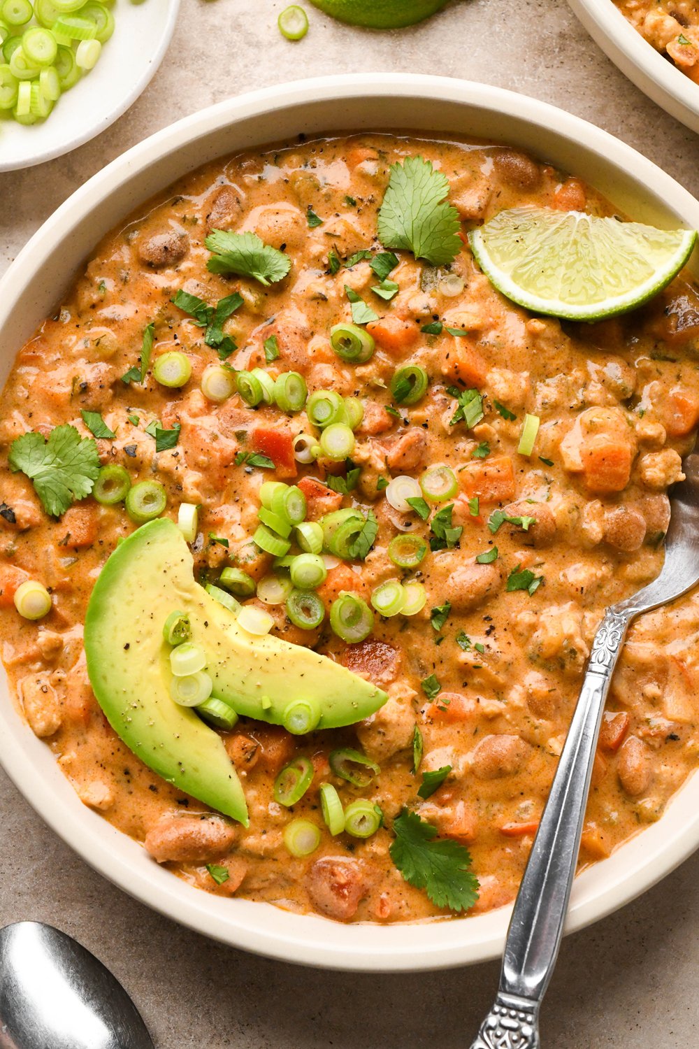 A hearty-looking bowl of creamy dairy free buffalo chicken chili garnished with sliced avocado, thinly sliced green onions, and chopped cilantro, with a spoon and lime wedge angled into the bowl.