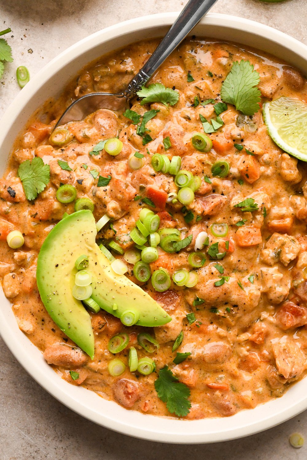 A bowl of creamy dairy free buffalo chicken chili garnished with sliced avocado, thinly sliced green onions, and chopped cilantro, with a spoon lifting out a bite of the thick and creamy soup.