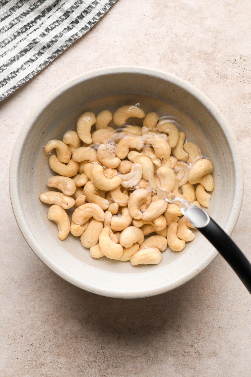 How to make Gluten Free Sausage Gravy: A tea kettle pouring just boiled water over raw cashews in a small ceramic heat-proof bowl.