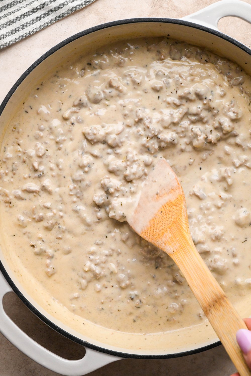 How to make Gluten Free Sausage Gravy: Cashew cream mixture in the skillet with cook sausage before simmering to thicken, a wooden spatula angled into the gravy to show the thick texture.