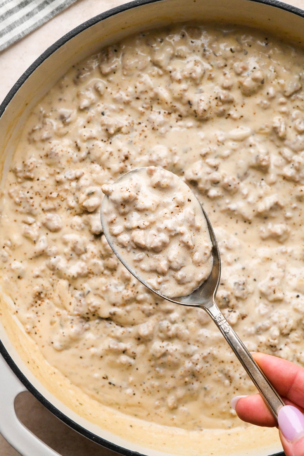How to make Gluten Free Sausage Gravy: Cashew cream mixture in the skillet with cook sausage before simmering to thicken, a large spoon lifting our a portion of the gravy to show the thick texture.