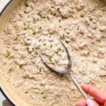 Thick and creamy gluten free sausage gravy in a wide shallow cream-colored ceramic skillet, garnished with black pepper and freshly snipped chives. A large antique metal spoon is lifting out a portion of the gravy to show the luscious texture.
