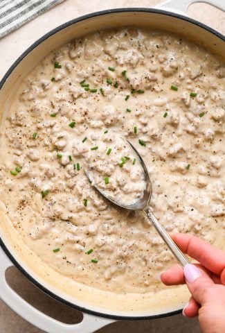 Thick and creamy gluten free sausage gravy in a wide shallow cream-colored ceramic skillet, garnished with black pepper and freshly snipped chives. A large antique metal spoon is lifting out a portion of the gravy to show the luscious texture.