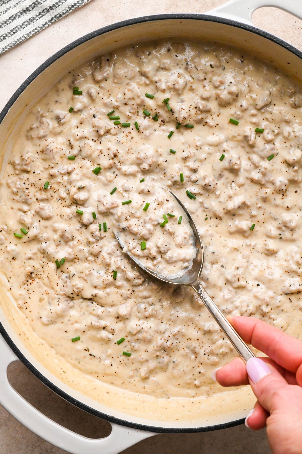 Thick and creamy gluten free sausage gravy in a wide shallow cream-colored ceramic skillet, garnished with black pepper and freshly snipped chives. A large antique metal spoon is lifting out a portion of the gravy to show the luscious texture.