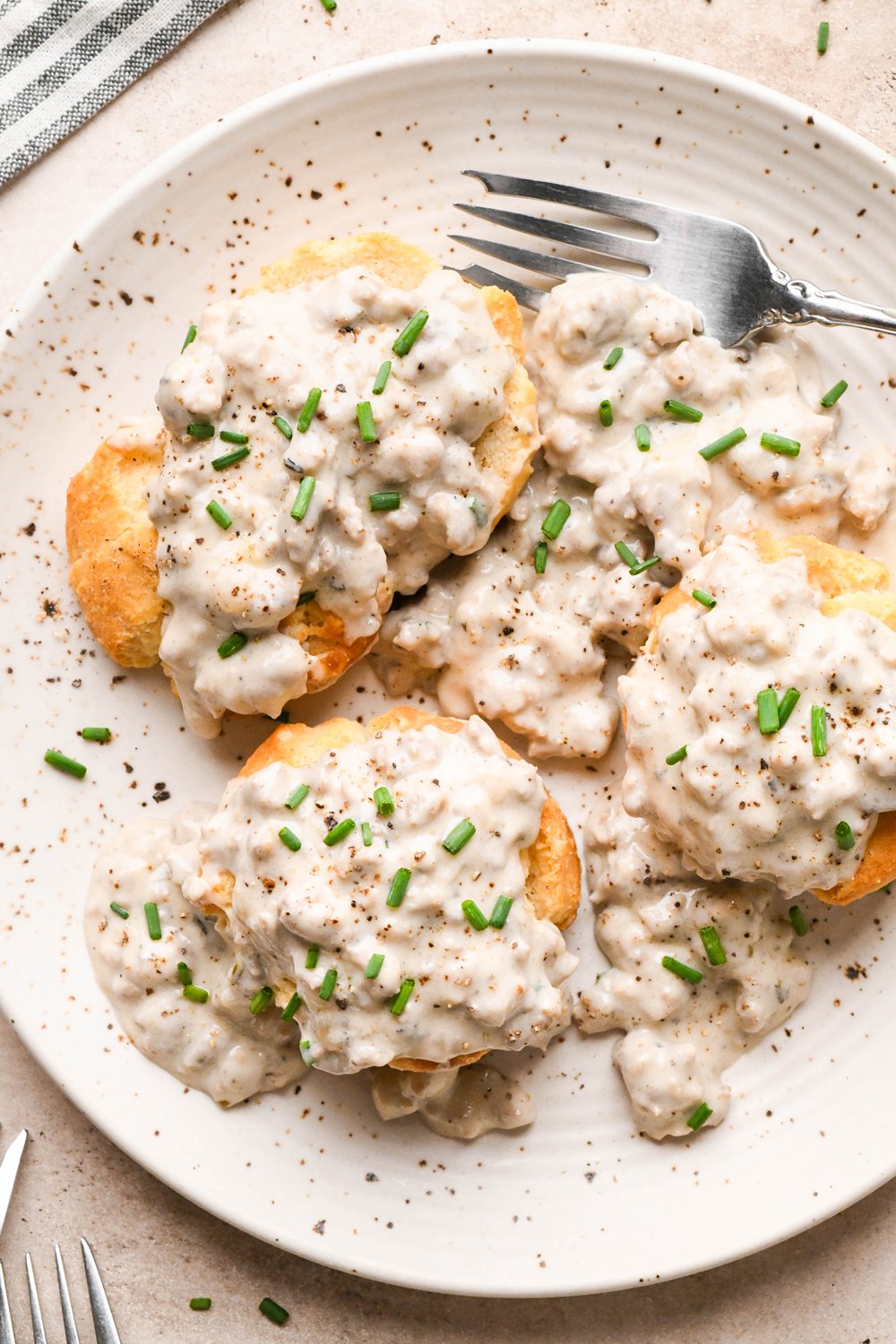 Gluten free sausage gravy on top of gluten free biscuits, garnished with black pepper and fresh chives on a cream colored speckled plate.