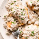 Gluten free sausage gravy on top of gluten free biscuits, garnished with black pepper and fresh chives on a cream colored speckled plate. A fork is on the edge of the plate, with a bite of the biscuit and gravy on it.