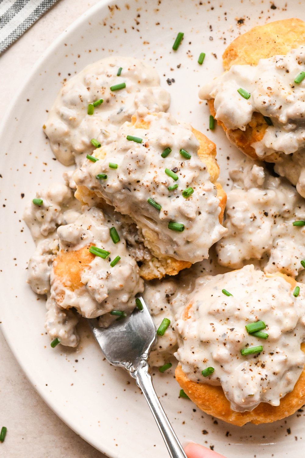 Gluten free sausage gravy on top of gluten free biscuits, garnished with black pepper and fresh chives on a cream colored speckled plate. A fork is on the edge of the plate, with a bite of the biscuit and gravy on it.
