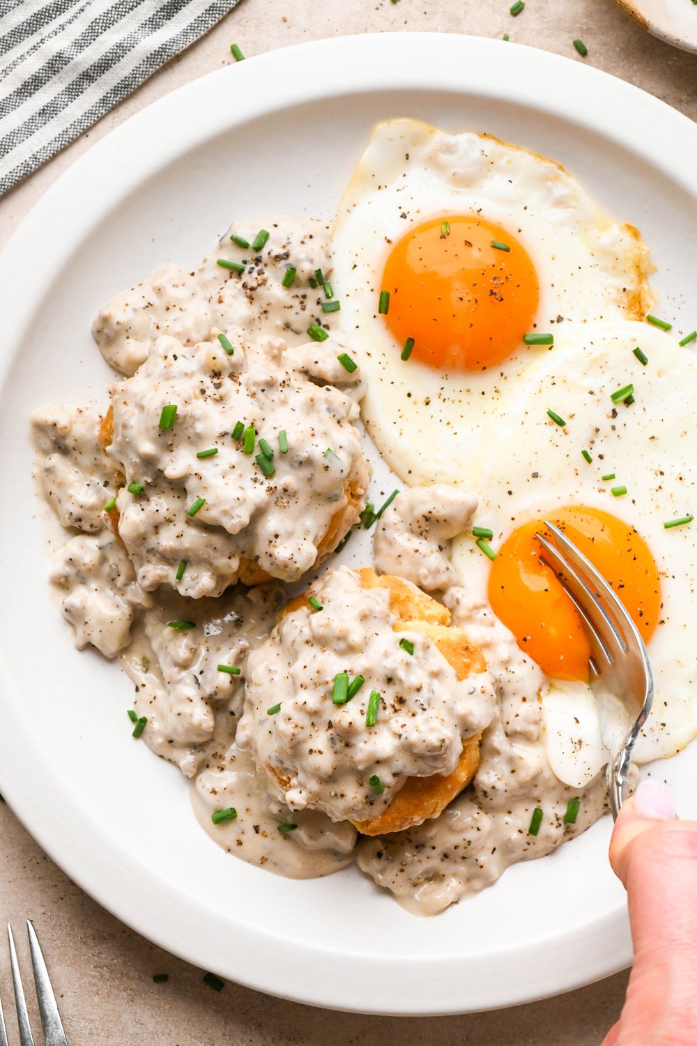 Creamy gluten free sausage gravy piled onto gluten free biscuits, next to two sunny side up eggs, and garnished with black pepper and fresh chives. A fork is cutting into the tender yolk of one of the fried eggs.