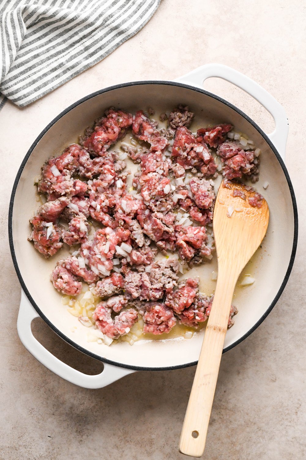 How to make Gluten Free Sausage Gravy: Breakfast sausage and diced onions in a large ceramic skillet during cooking, a spatula breaking up the meat into bite sized pieces.