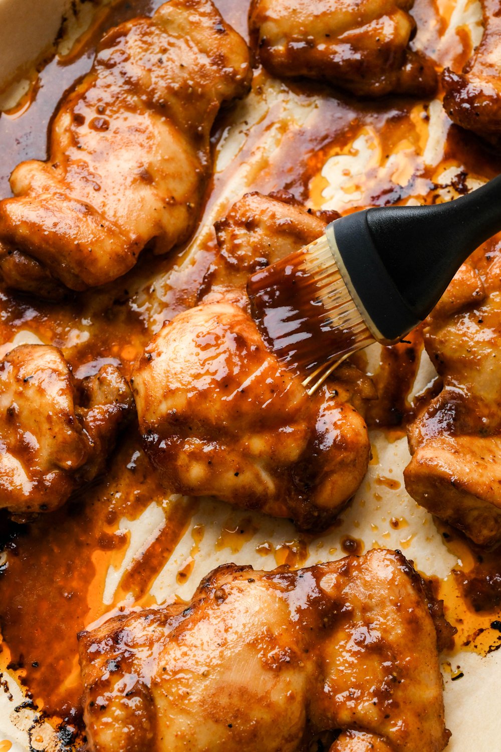 How to make BBQ Chicken Rice Bowls: Marinated chicken thighs on a baking sheet during cooking, a hand is brushing the partially cooked chicken with extra BBQ sauce.