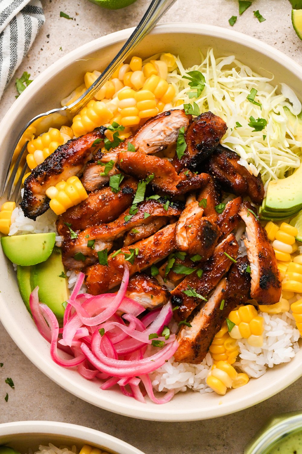 How to make BBQ Chicken Rice Bowls: Rice, shredded cabbage, cooked corn, avocado, pickled red onions, and thinly sliced BBQ chicken garnished with chopped cilantro in a shallow bowl.