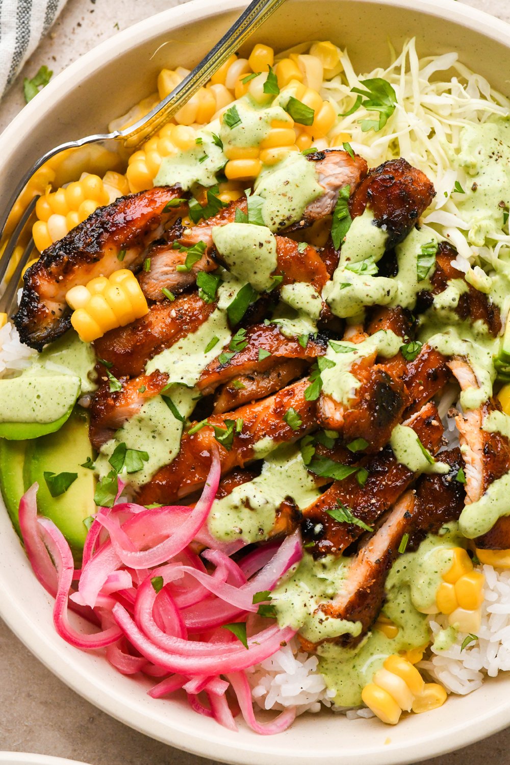 BBQ chicken rice bowl filled with rice, cooked sweet corn, thinly shredded cabbage, avocado, pickled red onions, and sliced BBQ chicken thighs. Drizzled with a bright green cilantro ranch dressing and garnished with chopped cilantro. 
