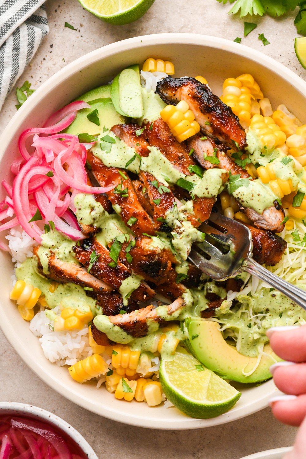 BBQ chicken rice bowl filled with rice, cooked sweet corn, thinly shredded cabbage, avocado, pickled red onions, and sliced BBQ chicken thighs. Drizzled with a bright green cilantro ranch dressing and garnished with chopped cilantro. A fork is digging into the chicken.