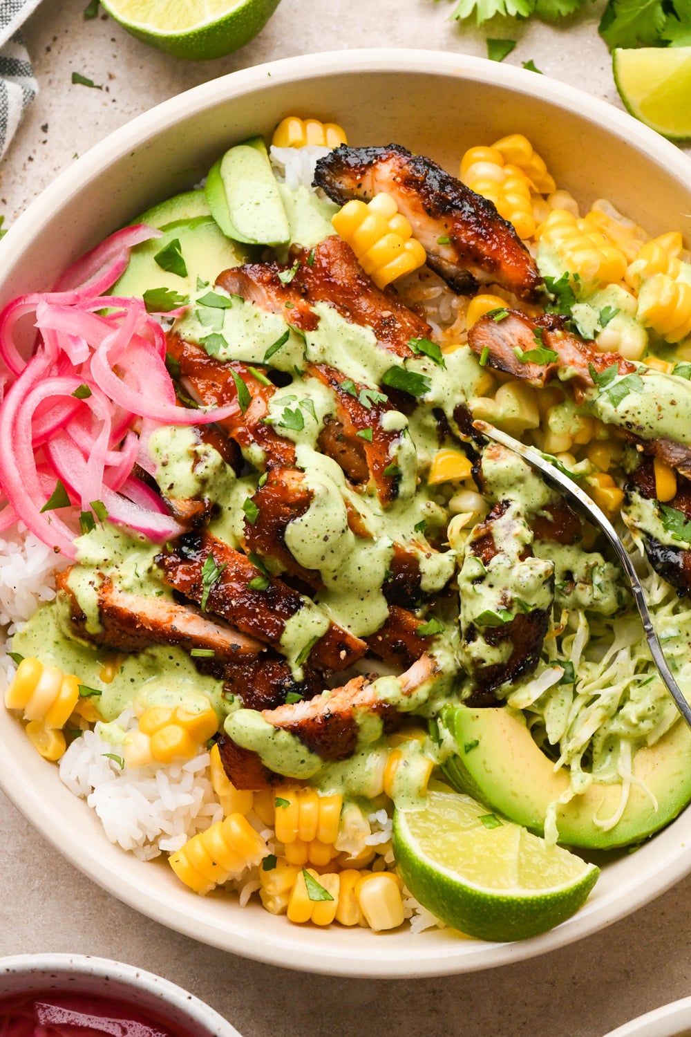 A bowl filled with sliced grilled chicken, corn, avocado, pickled onions, shredded lettuce, and rice, topped with a green sauce and garnished with lime wedges and cilantro. A fork rests in the bowl.
