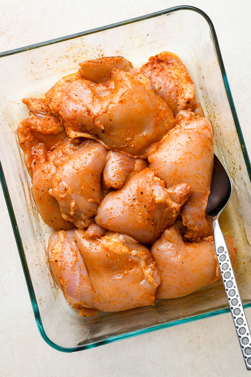 How to make BBQ Chicken Rice Bowls: Chicken thighs in a glass container, with spices and avocado oil coating the chicken.