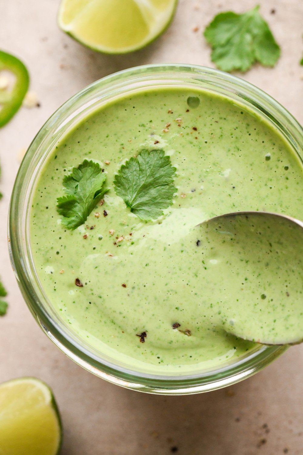A jar of thick and creamy bright green cilantro ranch dressing, with black pepper and a few cilantro leaves on top. A small spoon is dipping into the sauce to show the luscious texture.
