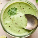 A jar of thick and creamy bright green cilantro ranch dressing, with black pepper and a few cilantro leaves on top. A small spoon is dipping into the sauce to show the luscious texture.