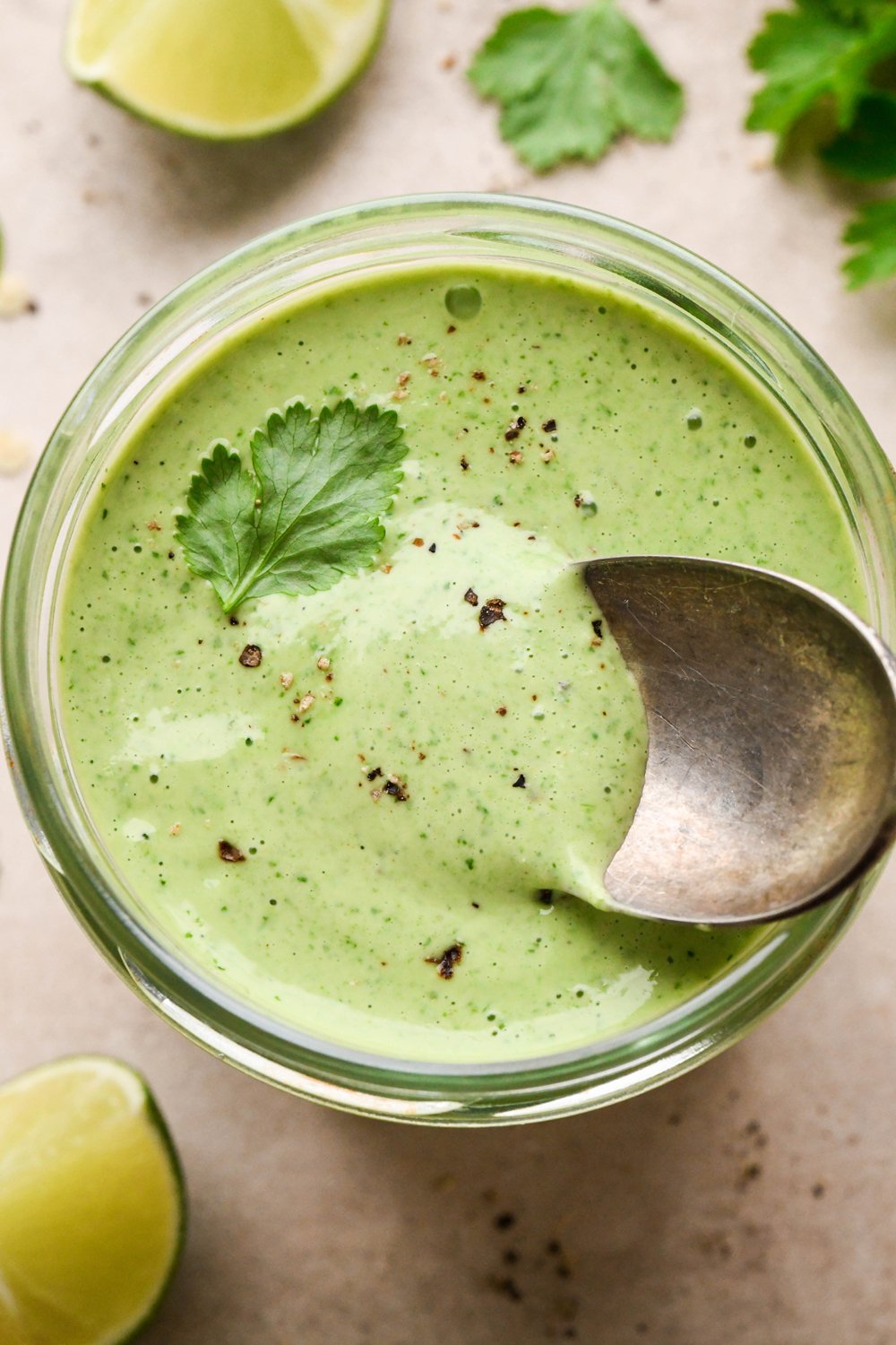 A jar of thick and creamy bright green cilantro ranch dressing, with black pepper and a few cilantro leaves on top. A small spoon is dipping into the sauce to show the luscious texture. 