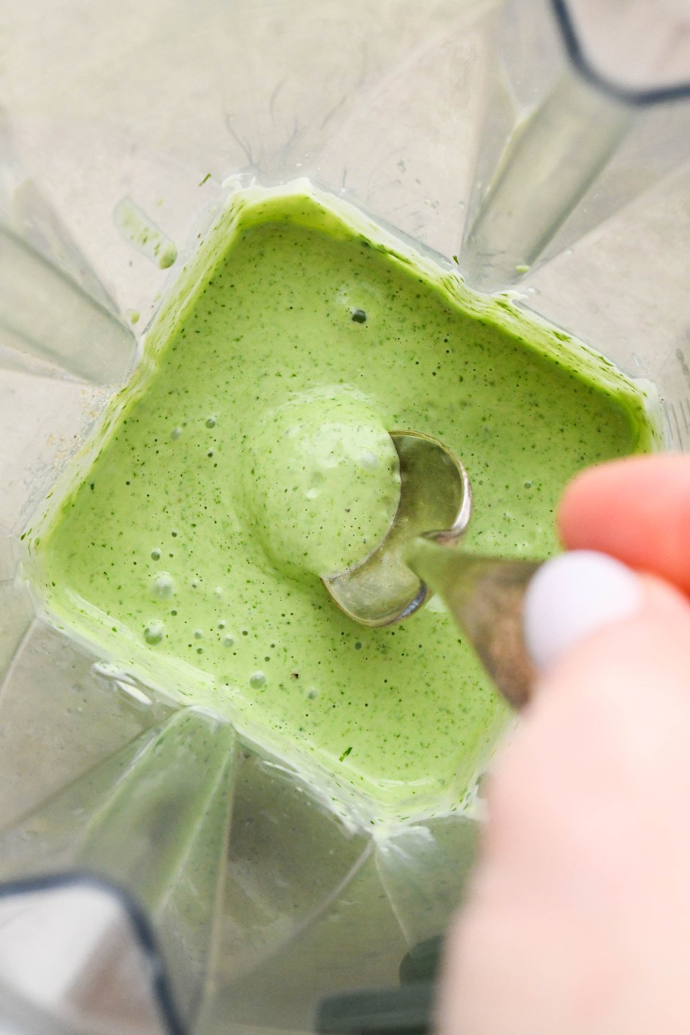 How to make Creamy 10-Minute Cilantro Ranch: A spoon dipping into the blender container with the process dressing, to show the thick and creamy texture.