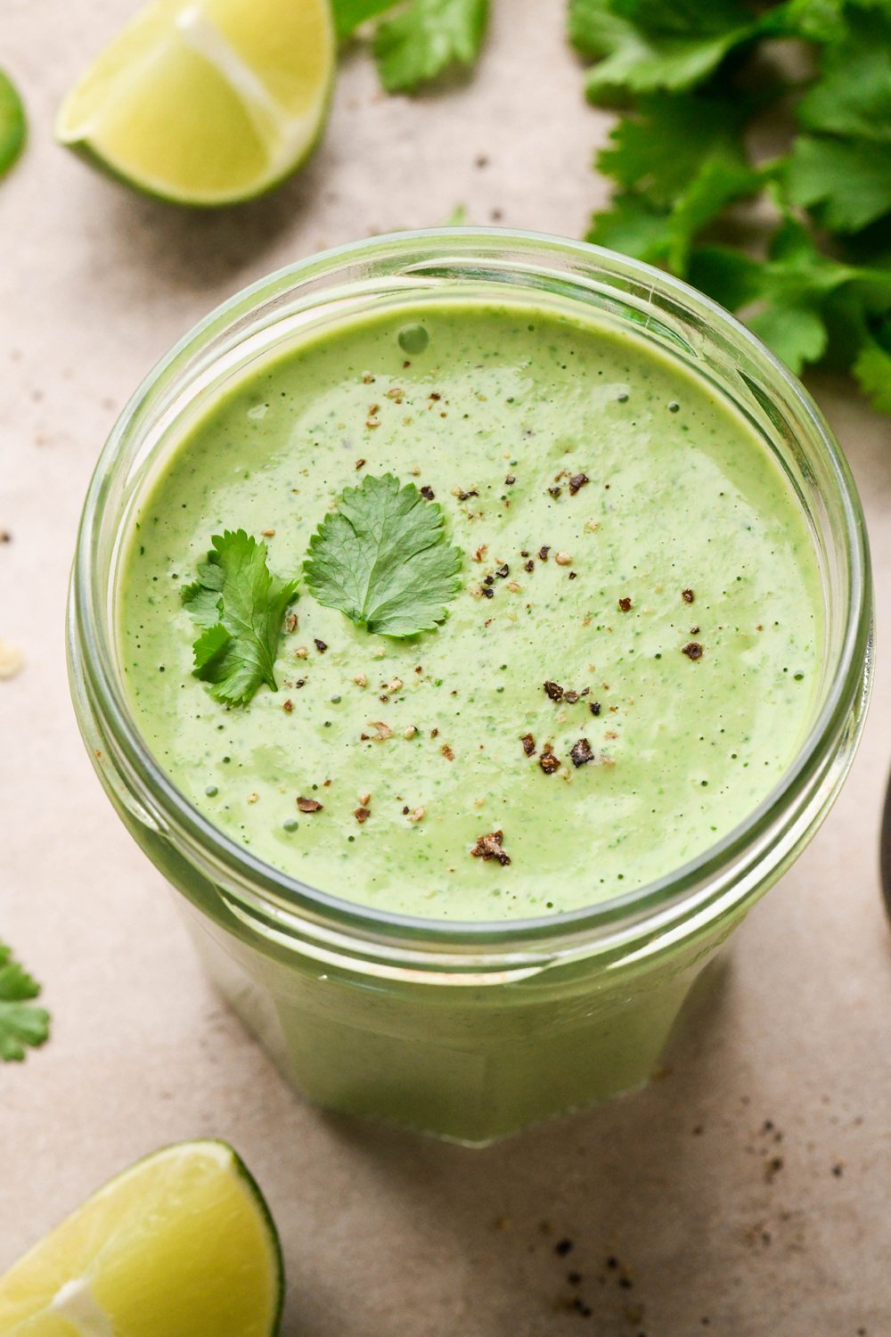 A jar of thick and creamy bright green cilantro ranch dressing, with black pepper and a few cilantro leaves on top. On a creamy light brown background, surrounded by some cut lime wedges and small bundle of fresh cilantro.