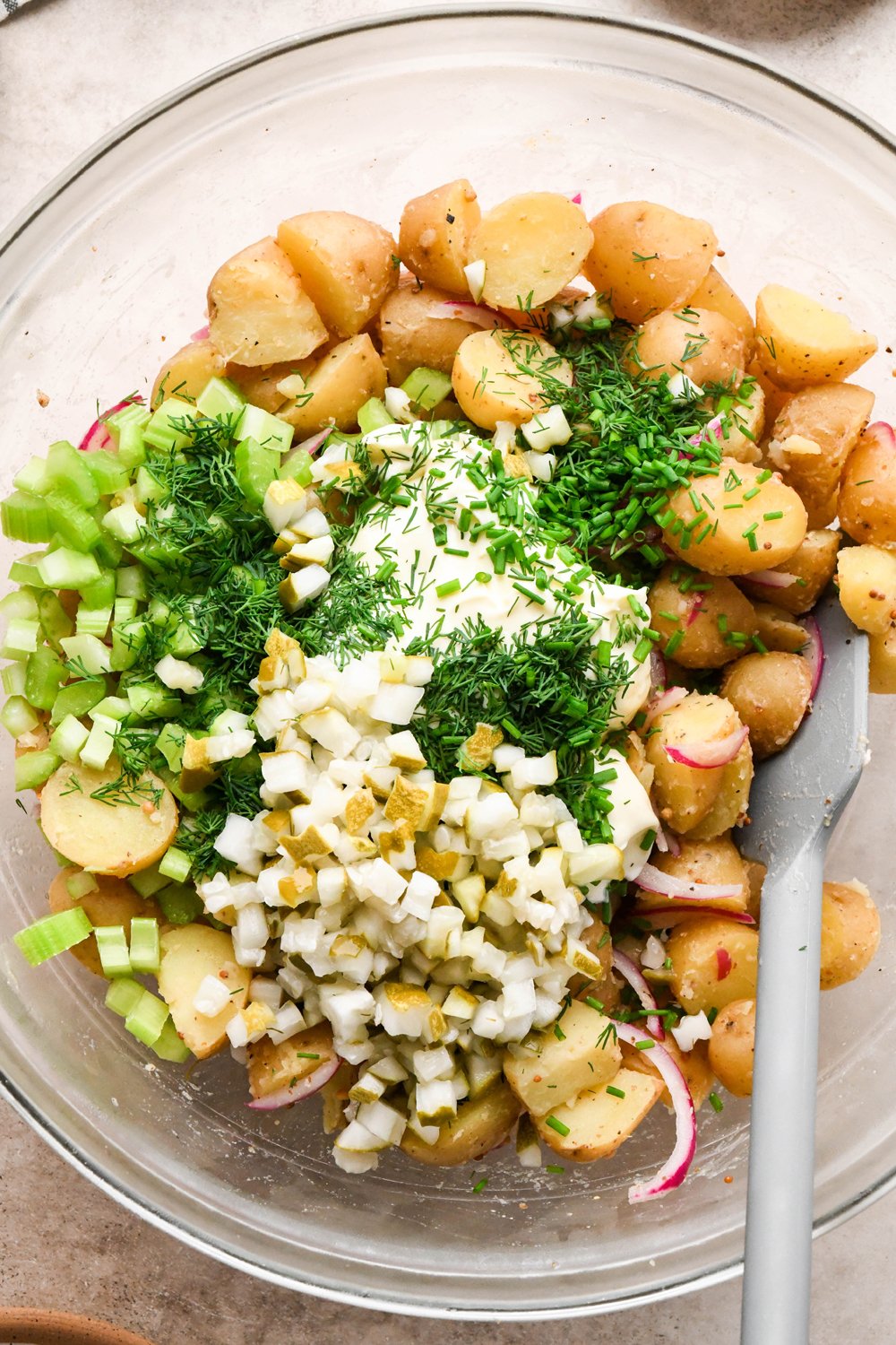 How to make Dill Pickle Potato Salad with Creamy Dill Dressing: Mayonnaise, chopped pickles, diced celery, and fresh herbs added to the large glass mixing bowl with potatoes, before stirring to combine.