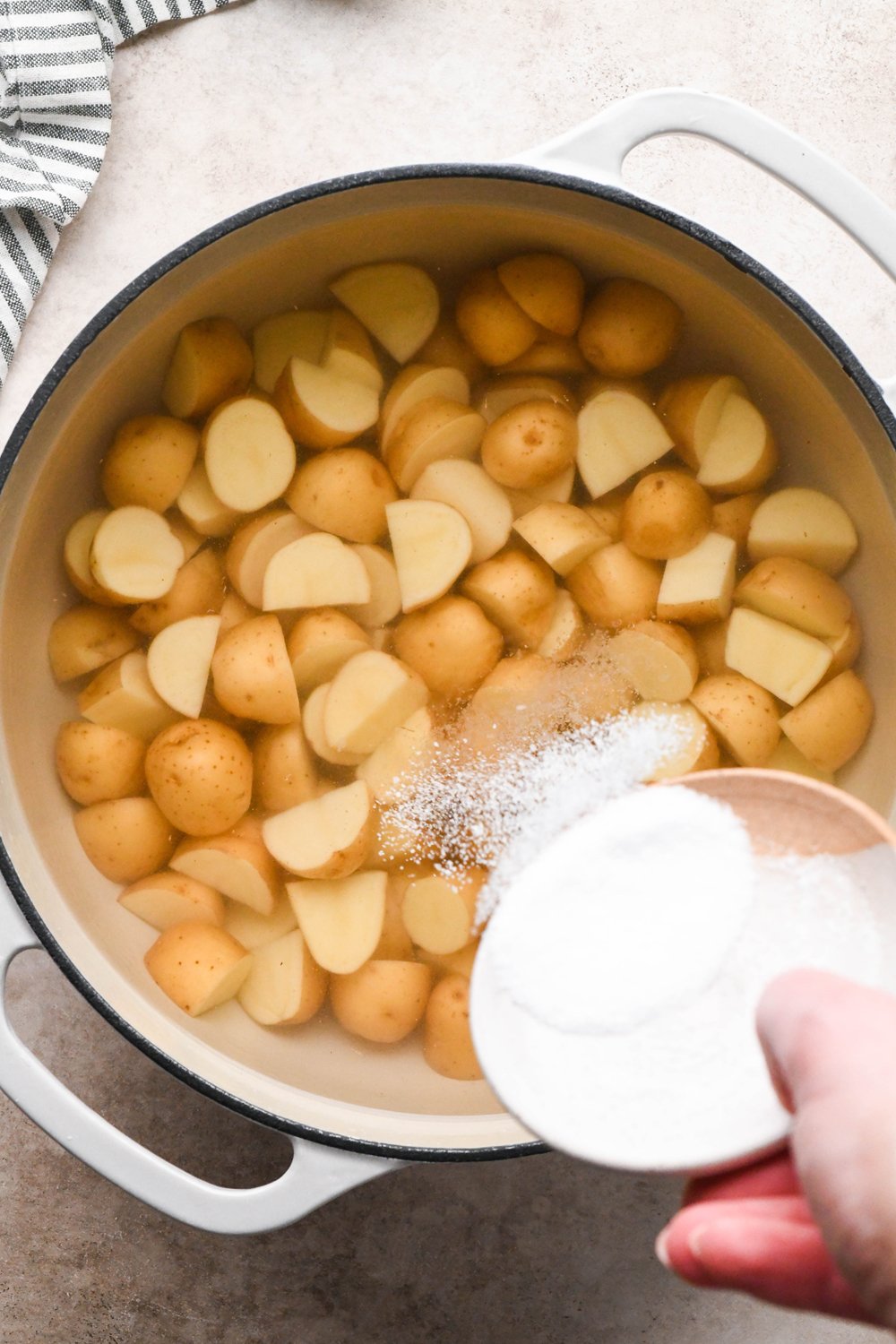 How to make Dill Pickle Potato Salad with Creamy Dill Dressing: A hand pouring kosher salt into a large pot that contains cut baby potatoes covered with water.
