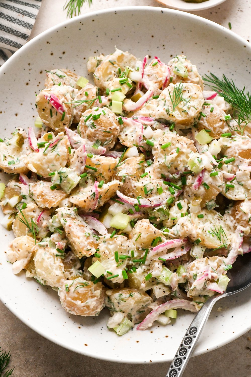 A large shallow speckled bowl filled with creamy mayo-based dill pickle potato salad. A spoon is angled into the bowl, and the potato salad is garnished with fresh dill, fresh snipped chives, and black pepper.